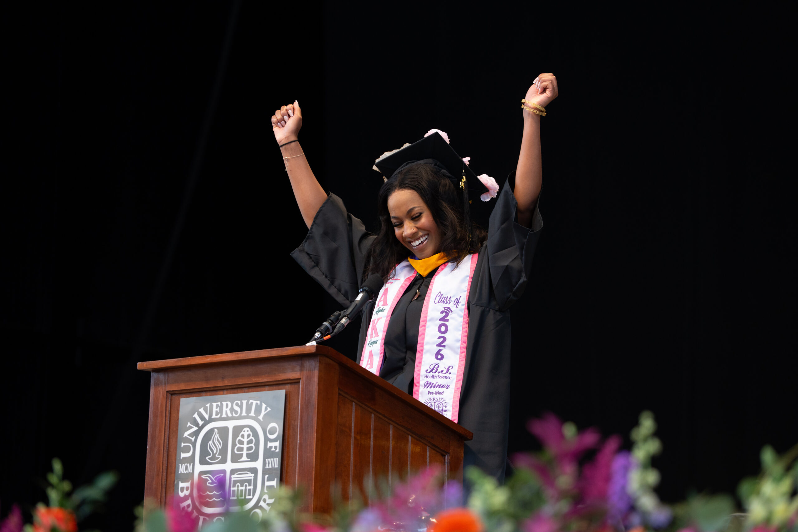 Woman in commencement regalia smiling at a podium with her hands raised in the air.