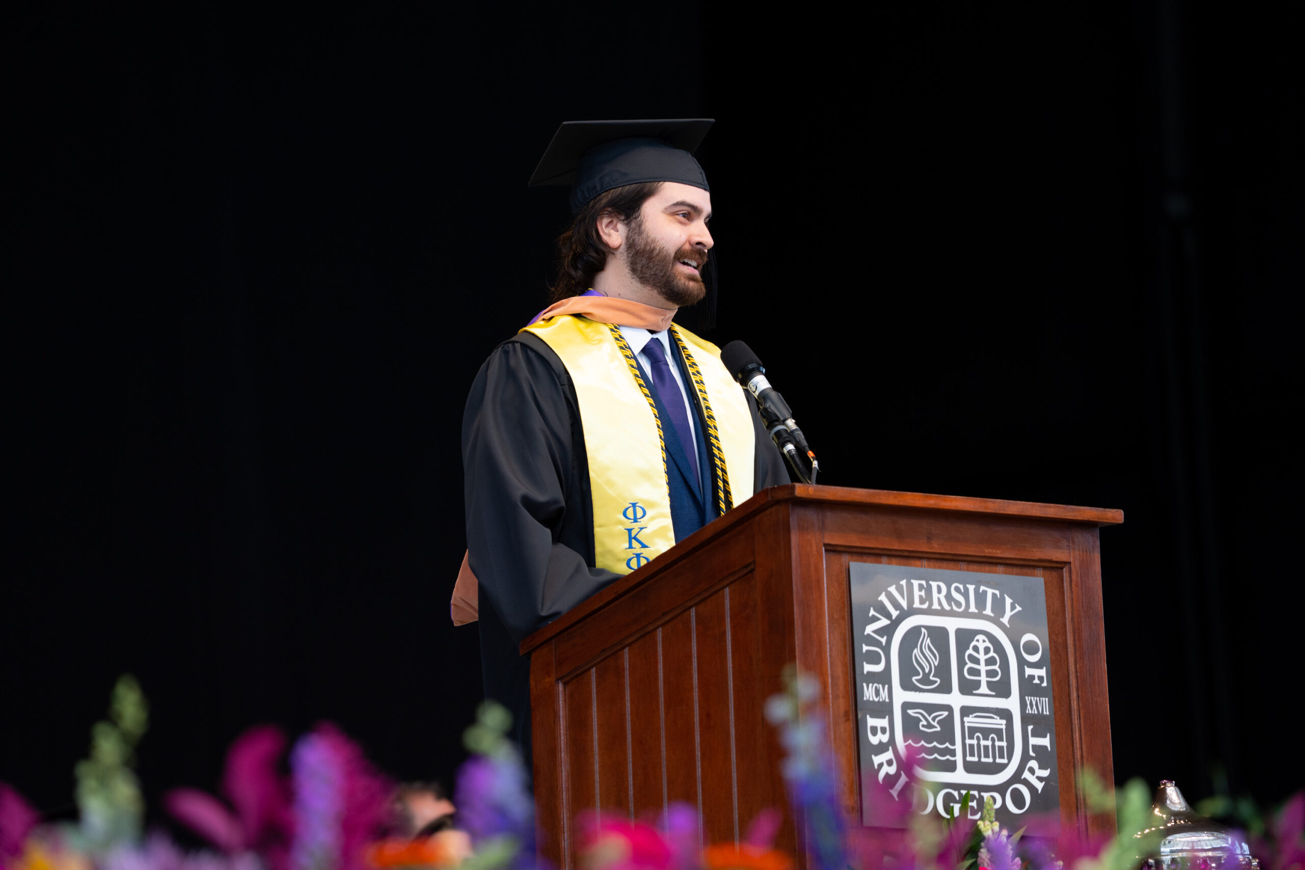 Man in commencement regalia standing at a podium smiling.