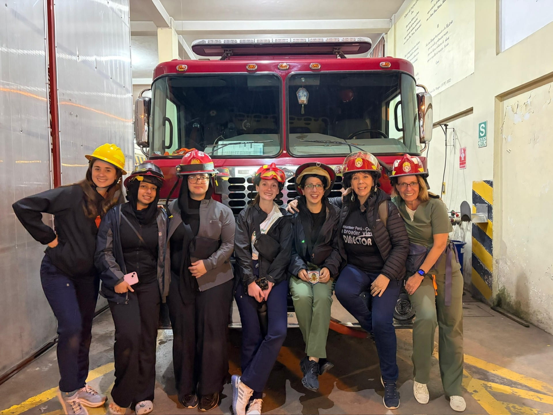 Seven women sitting or standing at the front of a firetruck wearing fire hats.