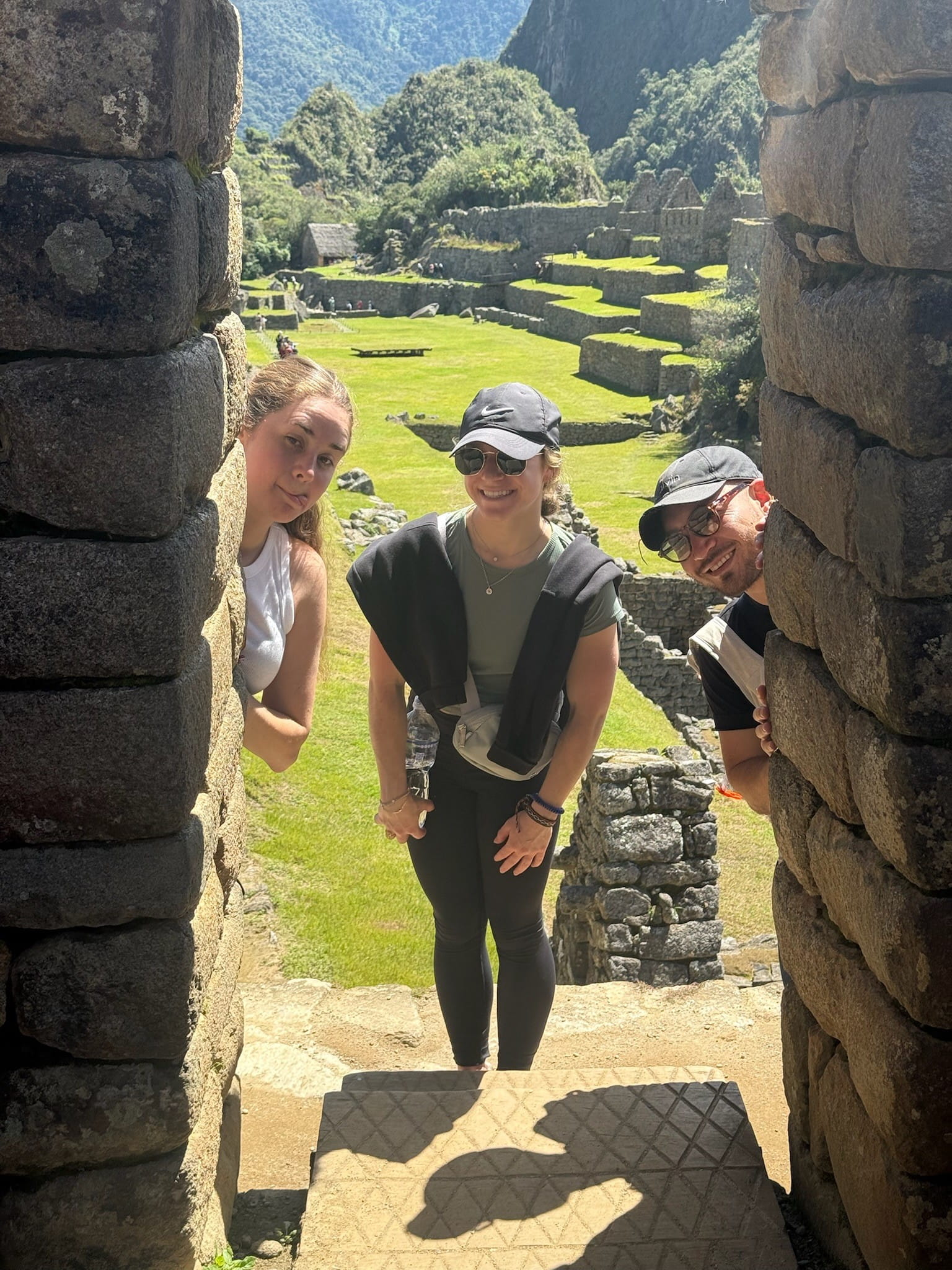 Two women and a man peeking through a stone opening with Machu Pichu in the background.