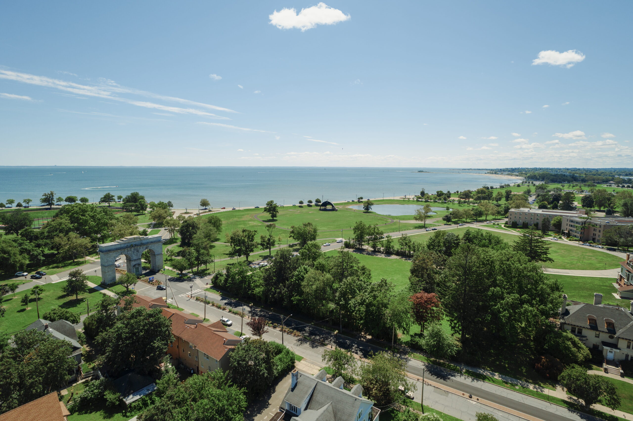 Aerial view of University of Bridgeport's campus, showing green open spaces, campus buildings, the Perry Memorial Arch, and the Long Island Sound shoreline on a clear, sunny day.