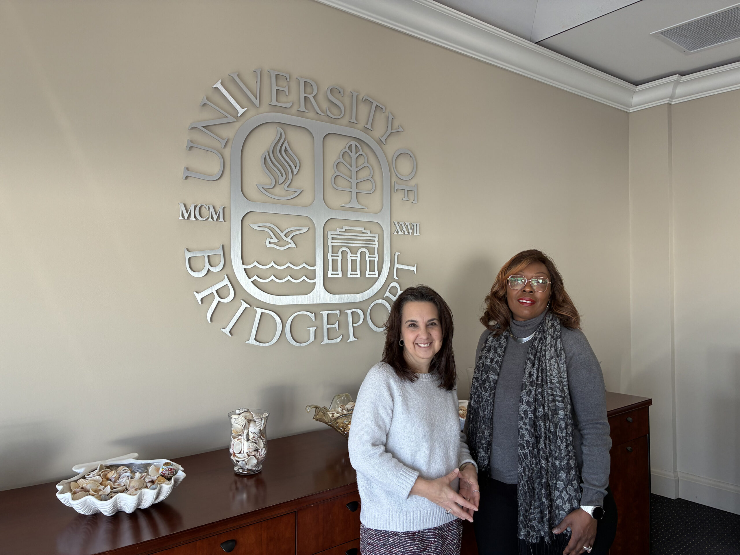 Two women standing in front of a University of Bridgeport logo posing for a photograph.