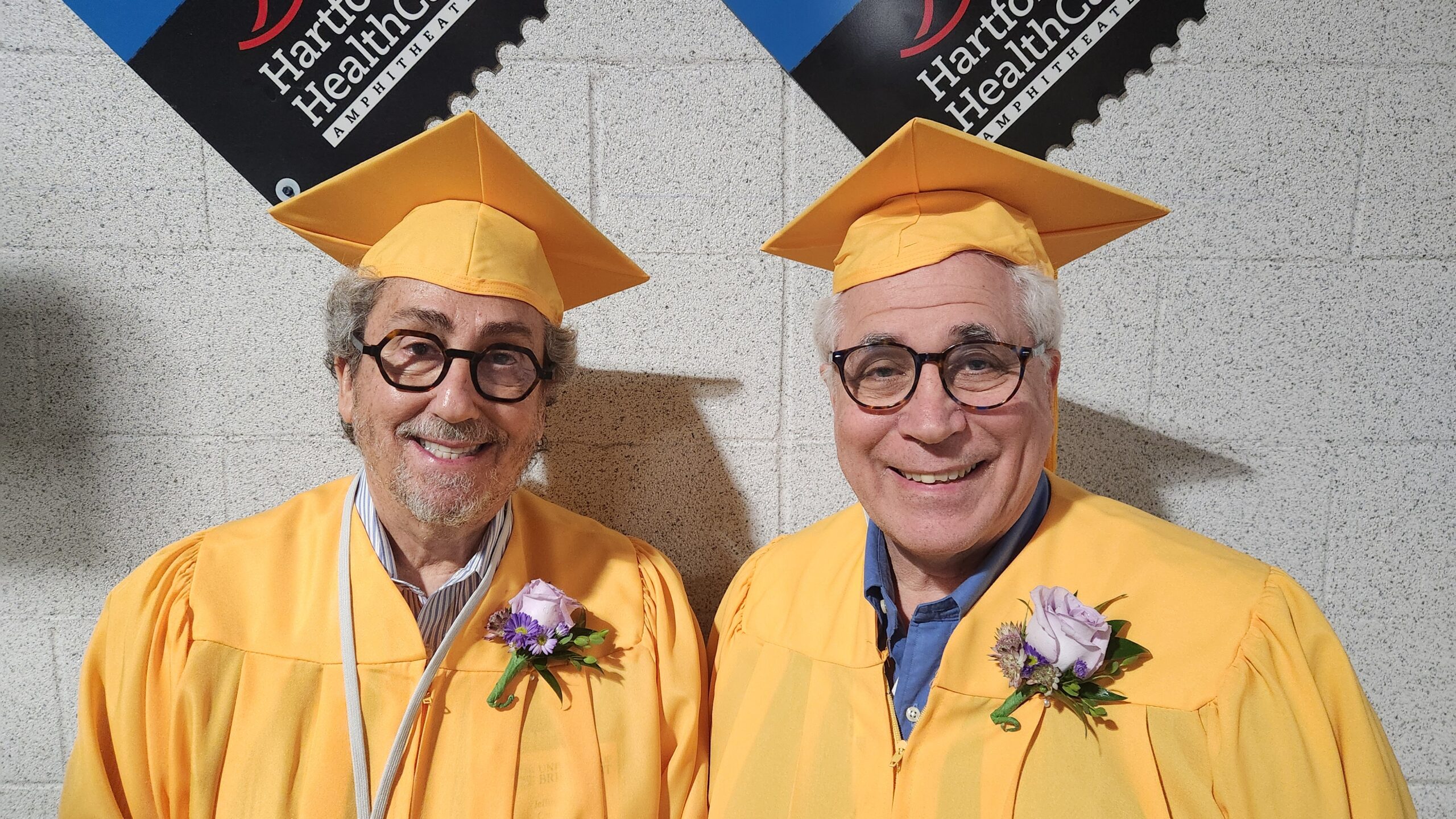 Close-up of Jeffrey Ostrove and David Rogers smiling at the camera in golden academic regalia, standing against a gray background before their induction as Golden Knights at University of Bridgeport’s 2025 Commencement ceremony.