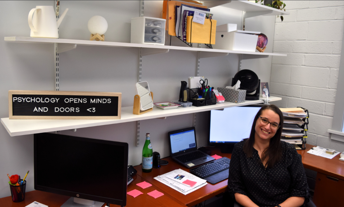 Professor poses in her office on the shelf if a sign that says, “psychology opens minds and doors.”