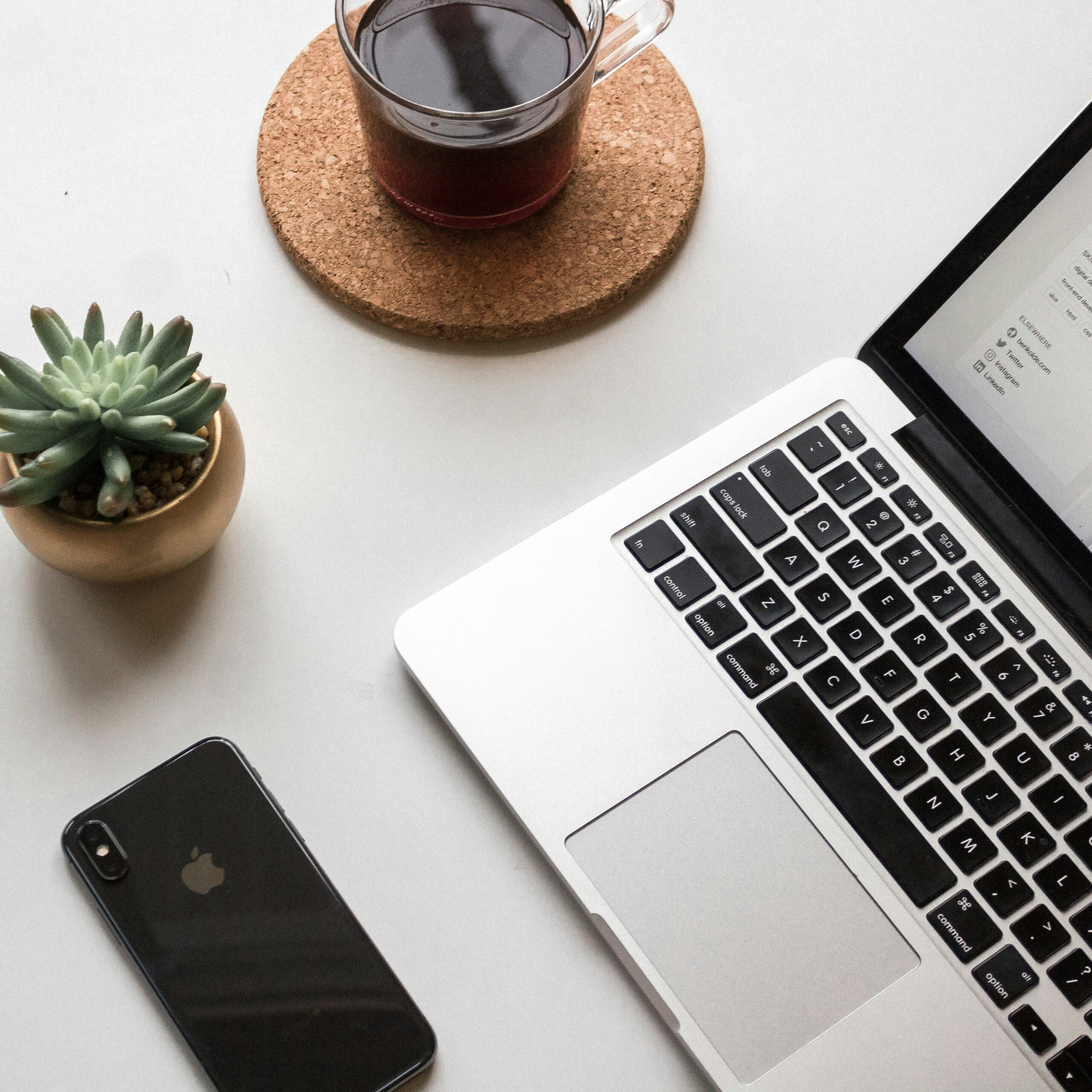 An open laptop on a white table with a black iPhone, a plant, and a cup of coffee.