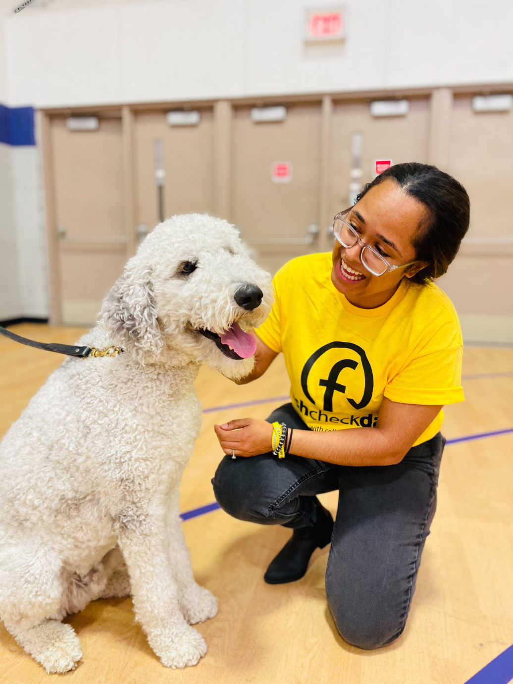 Close up of a woman wearing glasses and kneeling down petting a white dog who is sitting in front of her